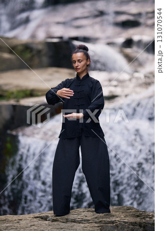 Woman practicing Tai Chi in nature, wearing a traditional black martial arts uniform, standing gracefully near a waterfall, focused on meditation and balance. Healthy lifestyle in asian culture 131070544
