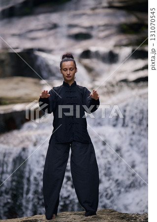 Woman practicing Tai Chi in nature, wearing a traditional black martial arts uniform, standing gracefully near a waterfall, focused on meditation and balance. Healthy lifestyle in asian culture Woman practicing Tai Chi in nature, wearing a traditional black martial arts uniform, standing gracefully near a waterfall, focused on meditation and balance. Healthy lifestyle in asian culture 131070545