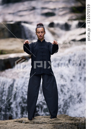 Woman practicing Tai Chi in nature, wearing a traditional black martial arts uniform, standing gracefully near a waterfall, focused on meditation and balance. Healthy lifestyle in asian culture Woman practicing Tai Chi in nature, wearing a traditional black martial arts uniform, standing gracefully near a waterfall, focused on meditation and balance. Healthy lifestyle in asian culture 131070559