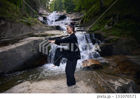 Woman practicing Tai Chi in nature, wearing a traditional black martial arts uniform, standing gracefully near a waterfall, focused on meditation and balance. Healthy lifestyle in asian culture 131070571