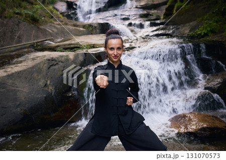 Woman practicing Tai Chi in nature, wearing a traditional black martial arts uniform, standing gracefully near a waterfall, focused on meditation and balance. Healthy lifestyle in asian culture 131070573