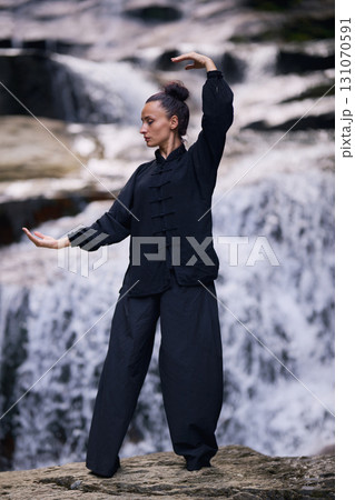 Woman practicing Tai Chi in nature, wearing a traditional black martial arts uniform, standing gracefully near a waterfall, focused on meditation and balance. Healthy lifestyle in asian culture Woman practicing Tai Chi in nature, wearing a traditional black martial arts uniform, standing gracefully near a waterfall, focused on meditation and balance. Healthy lifestyle in asian culture 131070591