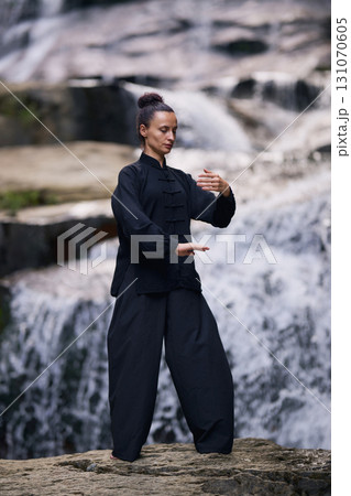 Woman practicing Tai Chi in nature, wearing a traditional black martial arts uniform, standing gracefully near a waterfall, focused on meditation and balance. Healthy lifestyle in asian culture 131070605