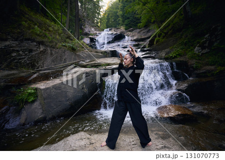 Woman practicing Tai Chi in nature, wearing a traditional black martial arts uniform, standing gracefully near a waterfall, focused on meditation and balance. Healthy lifestyle in asian culture Woman practicing Tai Chi in nature, wearing a traditional black martial arts uniform, standing gracefully near a waterfall, focused on meditation and balance. Healthy lifestyle in asian culture 131070773