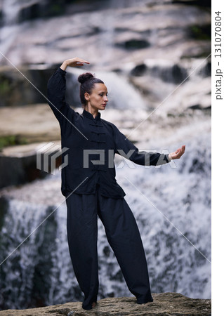 Woman practicing Tai Chi in nature, wearing a traditional black martial arts uniform, standing gracefully near a waterfall, focused on meditation and balance. Healthy lifestyle in asian culture Woman practicing Tai Chi in nature, wearing a traditional black martial arts uniform, standing gracefully near a waterfall, focused on meditation and balance. Healthy lifestyle in asian culture 131070804