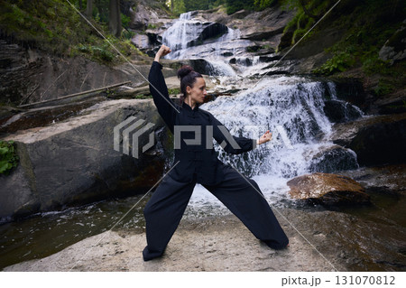 Woman practicing Tai Chi in nature, wearing a traditional black martial arts uniform, standing gracefully near a waterfall, focused on meditation and balance. Healthy lifestyle in asian culture 131070812