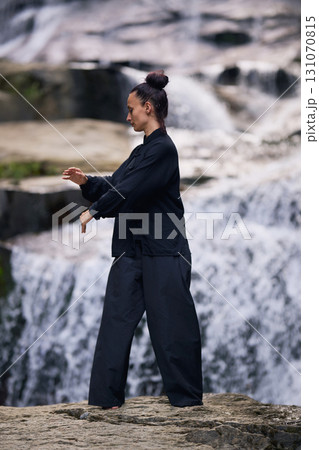 Woman practicing Tai Chi in nature, wearing a traditional black martial arts uniform, standing gracefully near a waterfall, focused on meditation and balance. Healthy lifestyle in asian culture 131070815