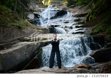 Beautiful young woman performing slow tai chi movements near a waterfall and river at sunset capturing harmony balance and fitness in nature. 131070819