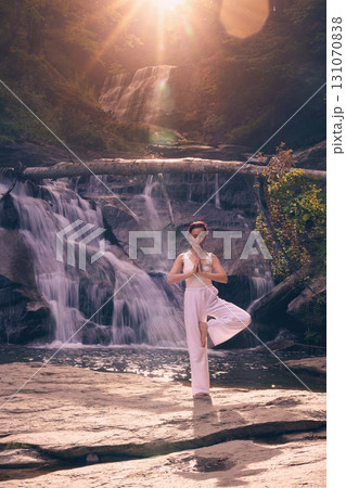 Woman doing yoga tree pose in front of waterfall at sunrise peaceful meditation in nature Woman doing yoga tree pose in front of waterfall at sunrise peaceful meditation in nature 131070838