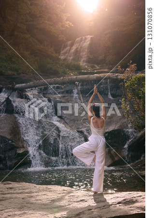 Woman doing yoga tree pose in front of waterfall at sunrise peaceful meditation in nature Woman doing yoga tree pose in front of waterfall at sunrise peaceful meditation in nature 131070856