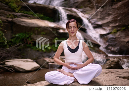 Young woman sitting in lotus pose under tropical waterfall meditating with calm strength and mindfulness fully connected to nature energy tranquility and inner balance 131070908