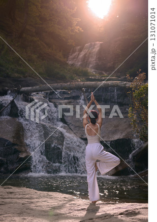 Woman doing yoga tree pose in front of waterfall at sunrise peaceful meditation in nature Woman doing yoga tree pose in front of waterfall at sunrise peaceful meditation in nature 131071014