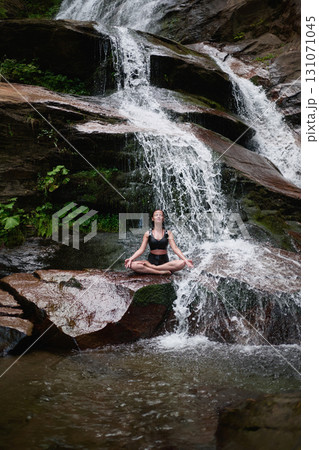 Young woman sitting in lotus pose under tropical waterfall meditating with calm strength and mindfulness fully connected to nature energy tranquility and inner balance Young woman sitting in lotus pose under tropical waterfall meditating with calm strength and mindfulness fully connected to nature energy tranquility and inner balance 131071045