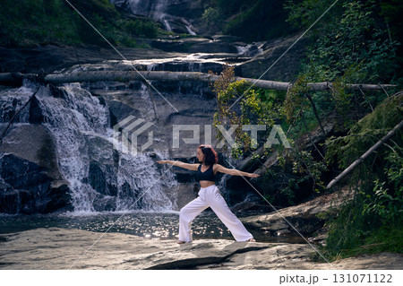 Woman practicing yoga warrior pose near waterfall at sunrise peaceful meditation in nature outdoor fitness Woman practicing yoga warrior pose near waterfall at sunrise peaceful meditation in nature outdoor fitness 131071122