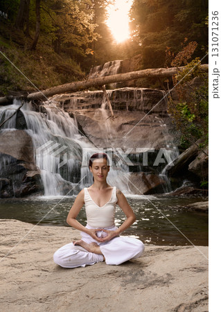 Young woman sitting in lotus pose under tropical waterfall meditating with calm strength and mindfulness fully connected to nature energy tranquility and inner balance 131071236
