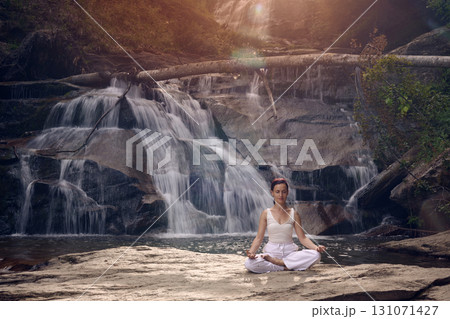 Young woman sitting in lotus pose under tropical waterfall meditating with calm strength and mindfulness fully connected to nature energy tranquility and inner balance 131071427