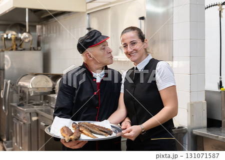 Two chefs in black uniforms working together while serving freshly fried fish in a commercial kitchen. Concept of seafood preparation, teamwork, culinary skills, and hospitality industry. Two chefs in black uniforms working together while serving freshly fried fish in a commercial kitchen. Concept of seafood preparation, teamwork, culinary skills, and hospitality industry. 131071587