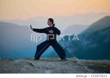 Woman practicing Tai Chi in traditional black outfit on mountain at sunrise, arms outstretched in meditation pose focusing on balance, energy, and mindfulness in nature. Woman practicing Tai Chi in traditional black outfit on mountain at sunrise, arms outstretched in meditation pose focusing on balance, energy, and mindfulness in nature. 131071621