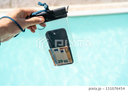 Hand holding waterproof transparent case with smartphone and euro banknotes against swimming pool background, concept of summer vacation safety, protection of valuables, travel security accessory. Hand holding waterproof transparent case with smartphone and euro banknotes against swimming pool background, concept of summer vacation safety, protection of valuables, travel security accessory. 131076454