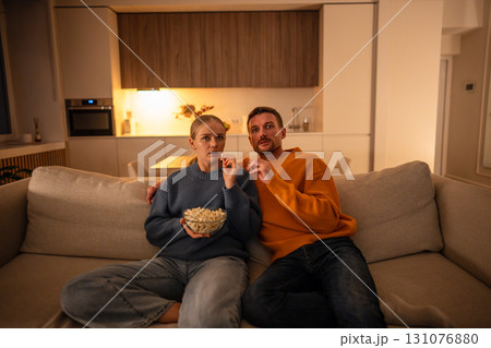Frightened couple watching horror movie with popcorn on sofa at home showing surprise and tension Frightened couple watching horror movie with popcorn on sofa at home showing surprise and tension 131076880