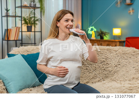 Pregnant woman enjoys dark chocolate rubs belly smiles calm lovely moment sweet treat at home sofa Pregnant woman enjoys dark chocolate rubs belly smiles calm lovely moment sweet treat at home sofa 131077067