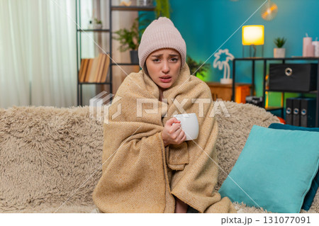 Young woman shivering on sofa under blanket wearing hat sipping hot tea hoping for gentle recovery 131077091