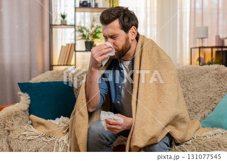 Young man sitting on home sofa, wrapped in a blanket, sneezing, reaching for tissues, feeling sick 131077545