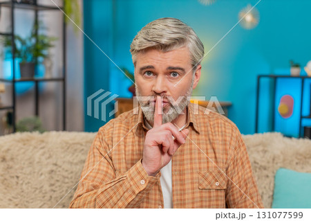 Mature man sitting on sofa making quiet gesture, expressing seriousness and secrecy at home 131077599