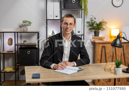 Young businessman looking to camera and writing down on diary in video chat at home office desk Young businessman looking to camera and writing down on diary in video chat at home office desk 131077980