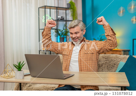 Mature man sitting at table with laptop, celebrating achievement win and victory in game at home Mature man sitting at table with laptop, celebrating achievement win and victory in game at home 131078012