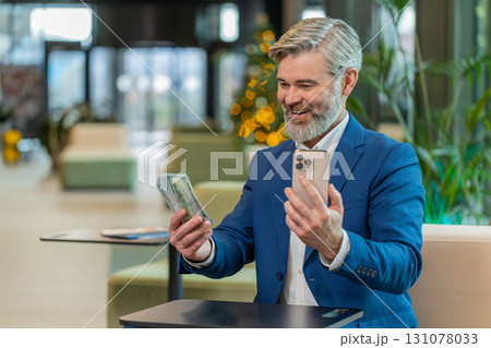 Successful businessman holding smartphone and counting dollar bill money fan sitting in office lobby 131078033