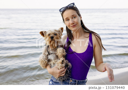 Happy young woman in swimsuit holding small dog at the beach. Authentic summer portrait with real emotions, Perfect for pet and travel themes. 131079440