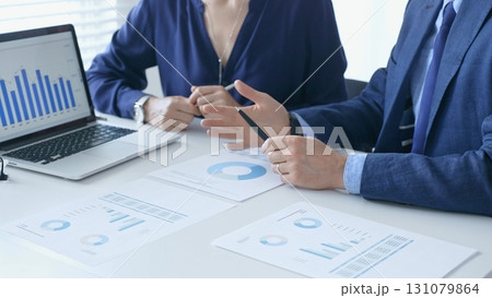 Businessman and businesswoman are analyzing financial data on a laptop and printed documents, pointing at charts and graphs during a meeting in bright office. Business people and finance concept 131079864