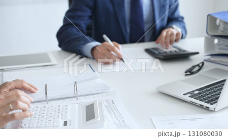 Two accountants are collaborating on a project, using a calculator and taking notes, surrounded by documents and technology in a busy office environment. Audit and taxes in business Two accountants are collaborating on a project, using a calculator and taking notes, surrounded by documents and technology in a busy office environment. Audit and taxes in business 131080030