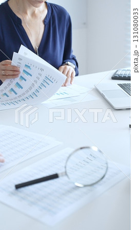 Female accountant reviewing financial documents, working collaboratively at white desk with blue folder stack, highlighting teamwork and business productivity. Audit and taxes in business Female accountant reviewing financial documents, working collaboratively at white desk with blue folder stack, highlighting teamwork and business productivity. Audit and taxes in business 131080033