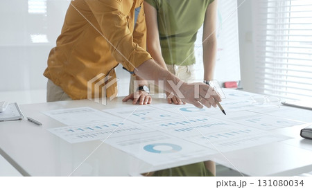 Two business professionals are examining financial reports, using a pen to point at specific data points on charts and graphs spread out on a white desk 131080034