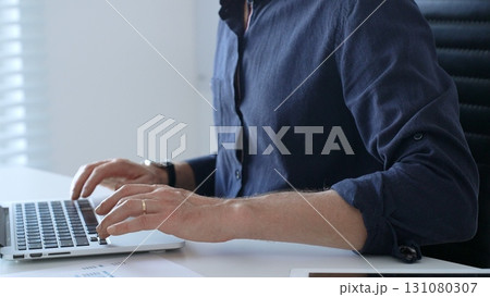 Businessman wearing a blue shirt is typing on a laptop with financial reports and a tablet on his desk, working diligently in a modern office environment. Business people concept 131080307