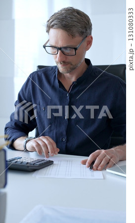 Businessman using a calculator and analyzing financial documents while preparing paperwork at an office desk. Audit and taxes, business concept Businessman using a calculator and analyzing financial documents while preparing paperwork at an office desk. Audit and taxes, business concept 131080333