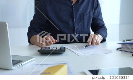 Businessman using a calculator and analyzing financial documents while preparing paperwork at an office desk. Audit and taxes, business concept Businessman using a calculator and analyzing financial documents while preparing paperwork at an office desk. Audit and taxes, business concept 131080334