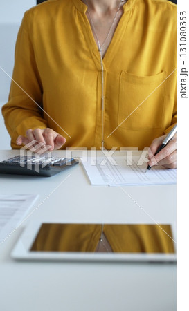 Businesswoman wearing yellow shirt sitting at desk and using calculator while taking notes, with laptop and folders in the background. Audit and taxes theme in business Businesswoman wearing yellow shirt sitting at desk and using calculator while taking notes, with laptop and folders in the background. Audit and taxes theme in business 131080353