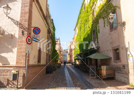 Cozy European alley lined with ivy-covered buildings and outdoor cafes. Sunlight filters through, highlighting rustic architecture and inviting dining setups. Strollers add life to the peaceful street 131081199
