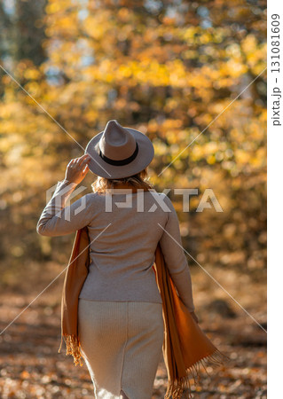 Woman Autumn Forest Walk: Back view of woman in hat and scarf strolling through autumnal forest; enjoying fall scenery. 131081609