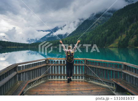 Woman with backpack standing on a wooden terrace with raised arms 131081637