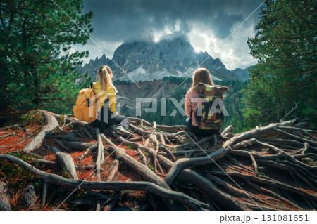 Two young women with backpacks sitting on tree roots in mountains 131081651