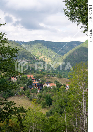 Peaceful rural village with houses surrounded by forested mountains under partly cloudy sky  131082761