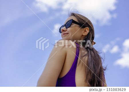 A woman smiles outdoors while wearing sunglasses in the summer. A happy smiled woman gazes into the distance She is wearing a purple swimsuit A woman smiles outdoors while wearing sunglasses in the summer. A happy smiled woman gazes into the distance She is wearing a purple swimsuit 131083629