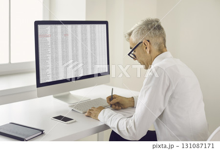 Senior man taking notes on paper while sitting at office desk with desktop computer Senior man taking notes on paper while sitting at office desk with desktop computer 131087171