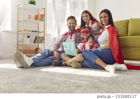 Happy family in red super hero costumes using laptop together sitting on the floor at home. 131088117