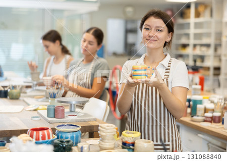 Girl stands in pottery workshop and shows created work, finished decorative vase - jug. 131088840