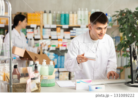 Portrait of young guy pharmacist standing at prescription counter and working on computer, stocktaking medicines at pharmacy shop and female assistant in background 131089222